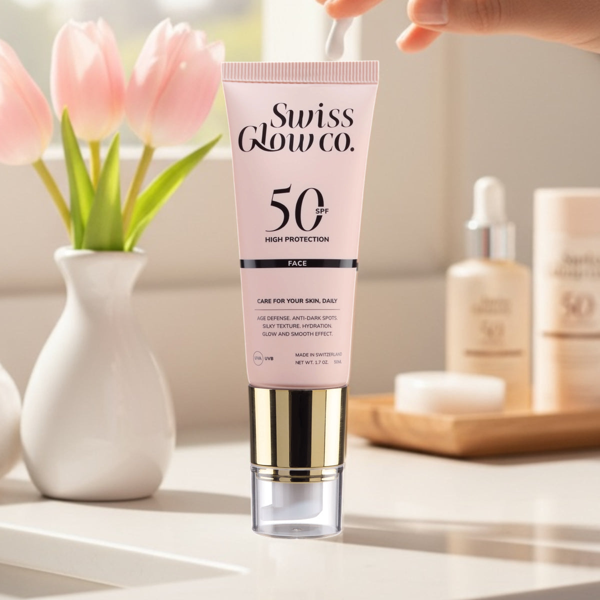 Swiss Glow Co. SPF50 Gesichtscreme-Tube auf Marmorablage neben rosa Tulpen, Hand drückt Creme aus der Tube